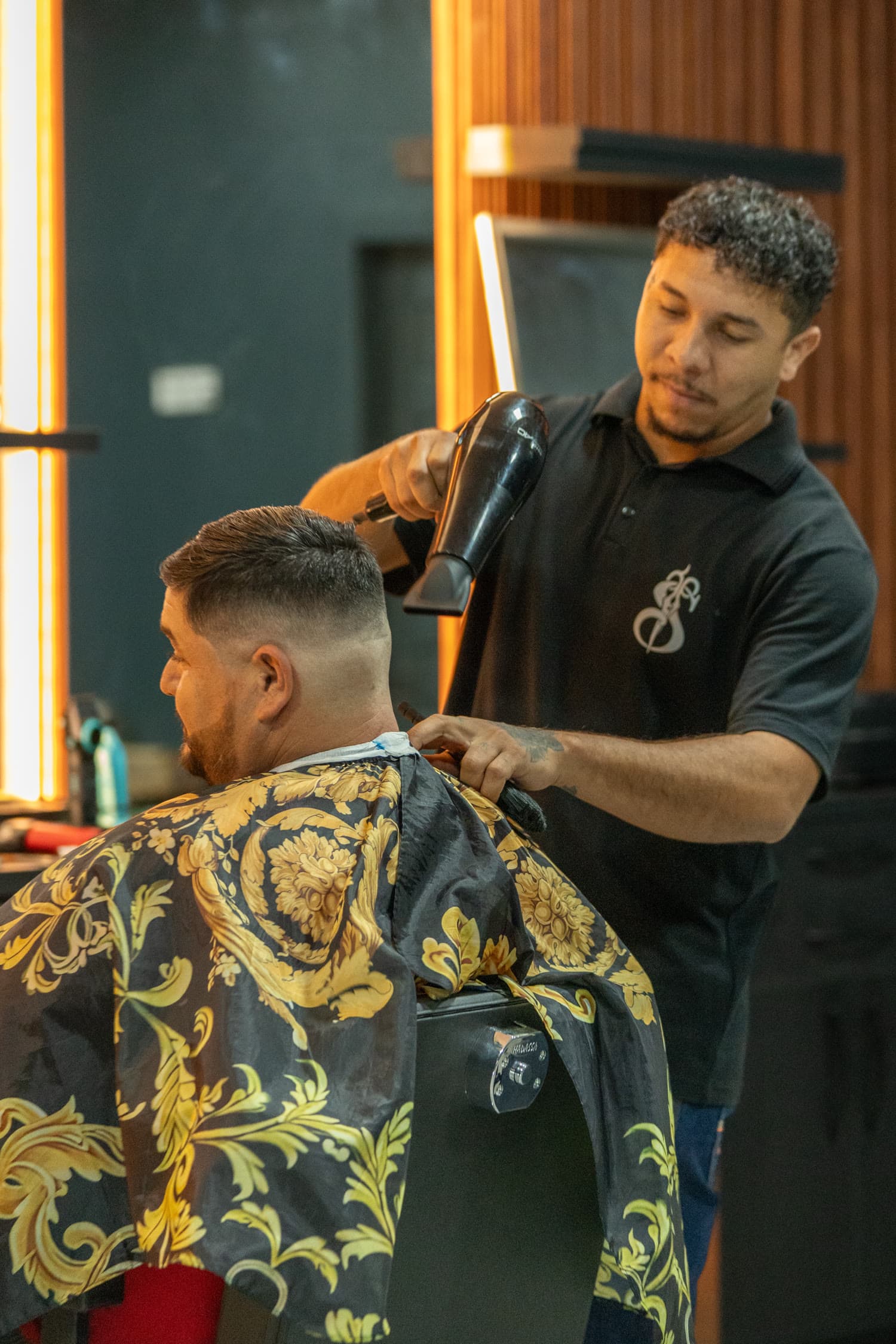 Barbeiro secando o cabelo de um cliente de capa dourada, ao lado dos painéis de LED